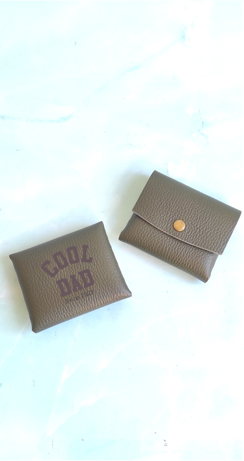 Dois porta-moedas castanhos em couro sintético, um com botão dourado e outro com texto 'COOL DAD'