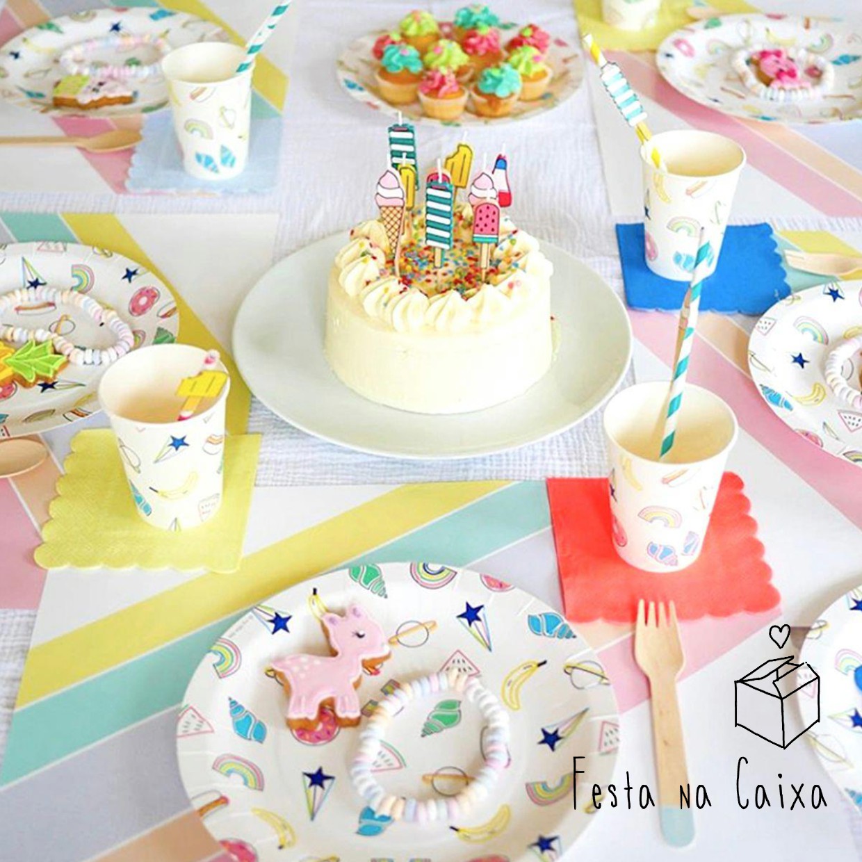 Mesa decorada para festa com pratos e copos coloridos, bolo branco com velas de aniversário, utensílios e padrão de unicórnios e estrelas