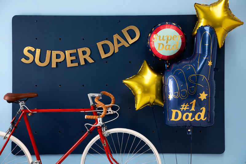 Decoração de festa 'Super Dad' com balões e letras douradas, painel azul escuro e bicicleta vermelha