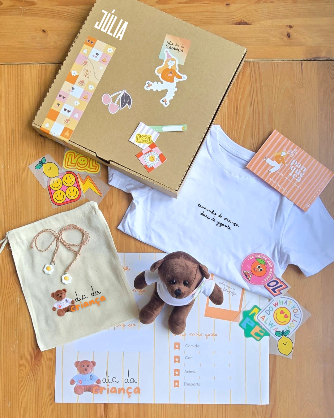 Kit de prendas com caixa de cartão, t-shirt branca, saquinho tecido, ursinho de peluche e autocolantes sobre mesa de madeira