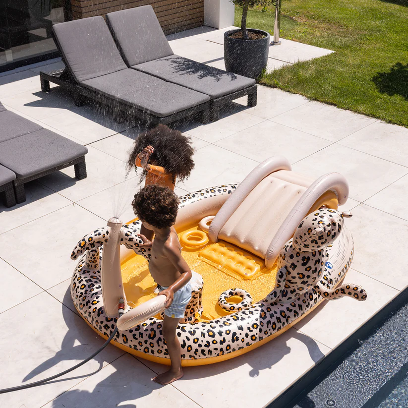 Piscina insuflável infantil com padrão de onça e escorregador ao lado de piscina e espreguiçadeiras