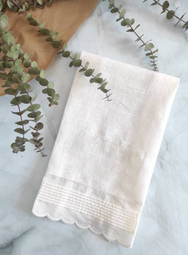 toalha branca bordada com detalhes e ramos verdes sobre fundo branco marmorizado
