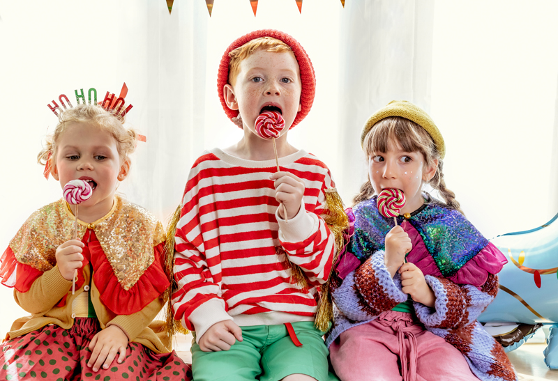 Três crianças a comer pirulitos, vestidos com roupas coloridas e festivas, com cortina branca atrás