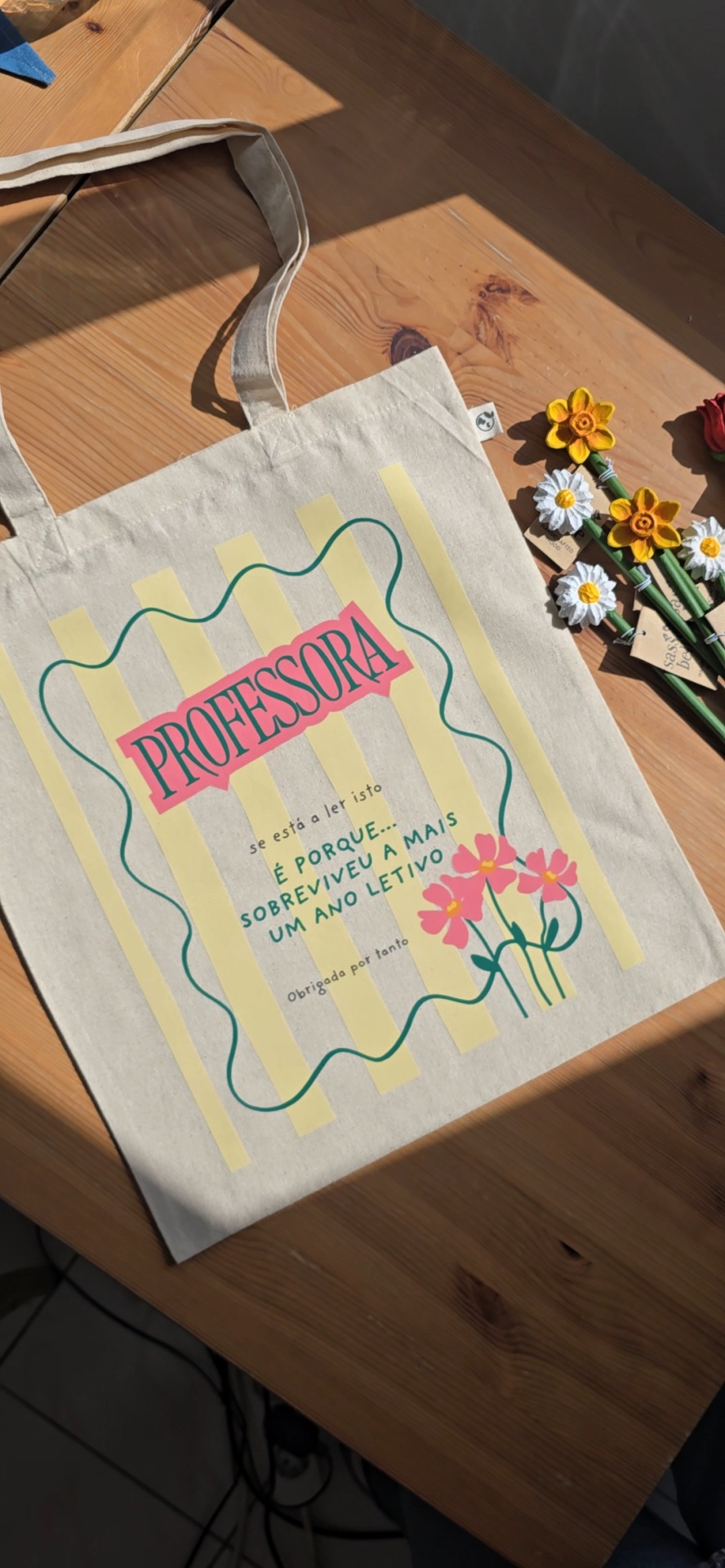 Mala de pano com estampa colorida e texto para professora, sobre mesa de madeira com flores artificiais ao lado.