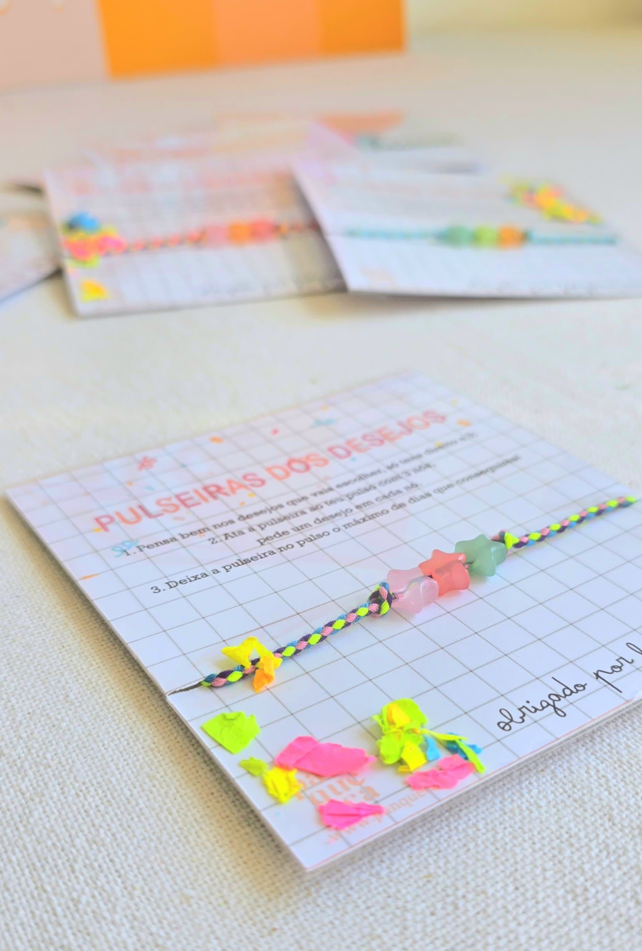 Pulseira de fios trançados coloridos com contas de estrela em cartão quadriculado com texto em português.