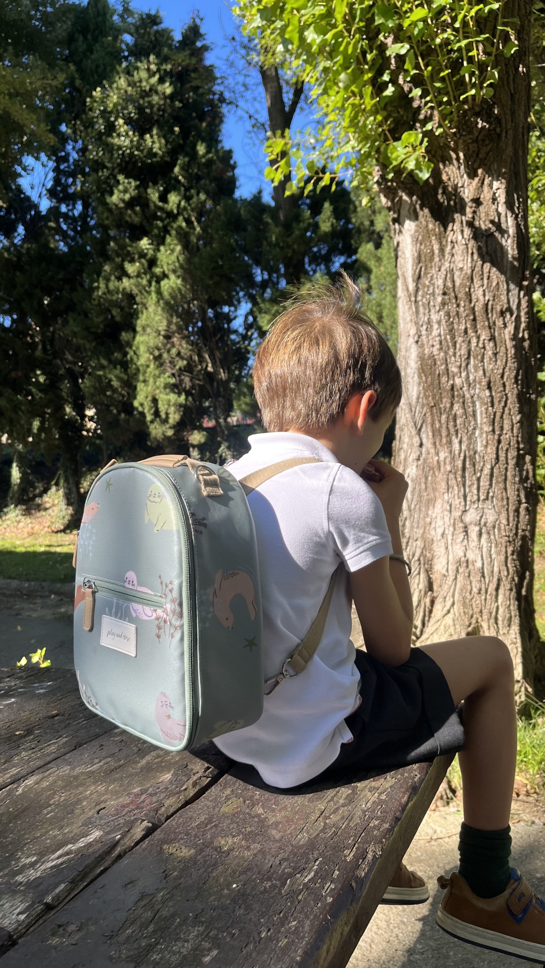 Criança sentada com mochila cinza claro com padrões coloridos em banco de madeira ao ar livre