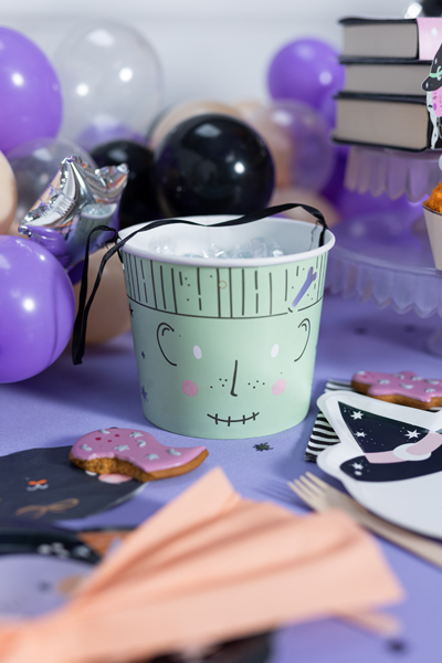 Balde de festa verde claro com rosto desenhado e alça preta entre decoração roxa e bolachas