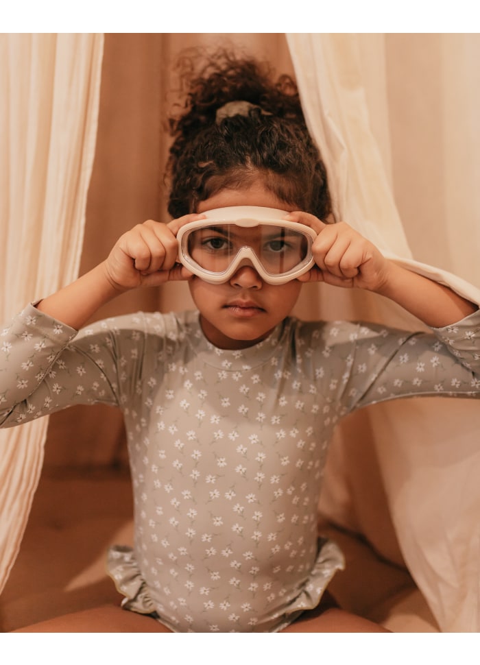 Criança com camisola cinzenta e óculos protetores brancos sentada entre tecidos.