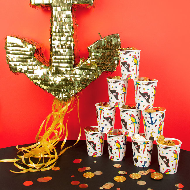 Copos descartáveis com padrões náuticos, piñata de âncora dourada e moedas de chocolate numa mesa preta com fundo vermelho