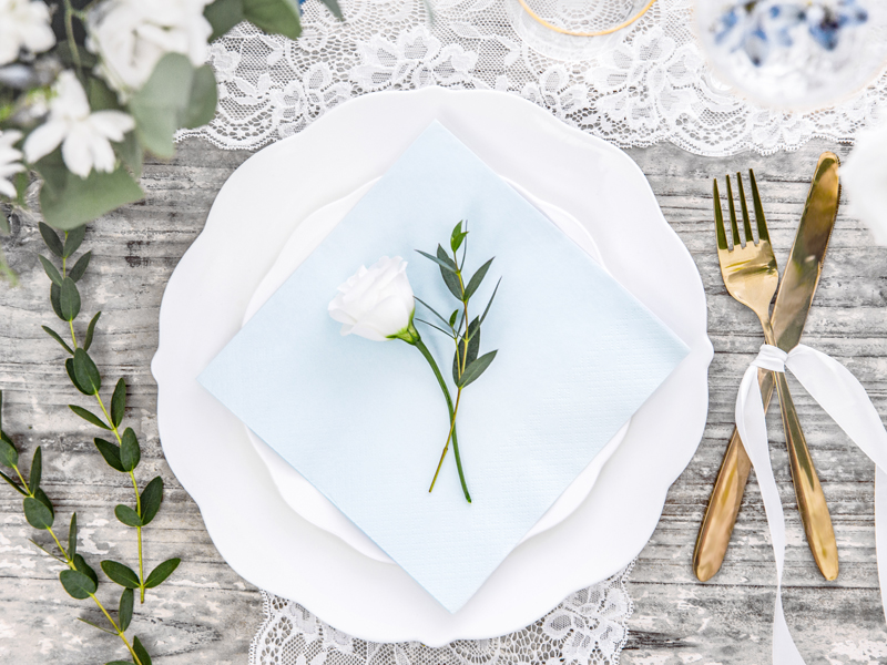 Mesa posta com prato branco, guardanapo azul claro e talheres dourados amarrados com fita branca