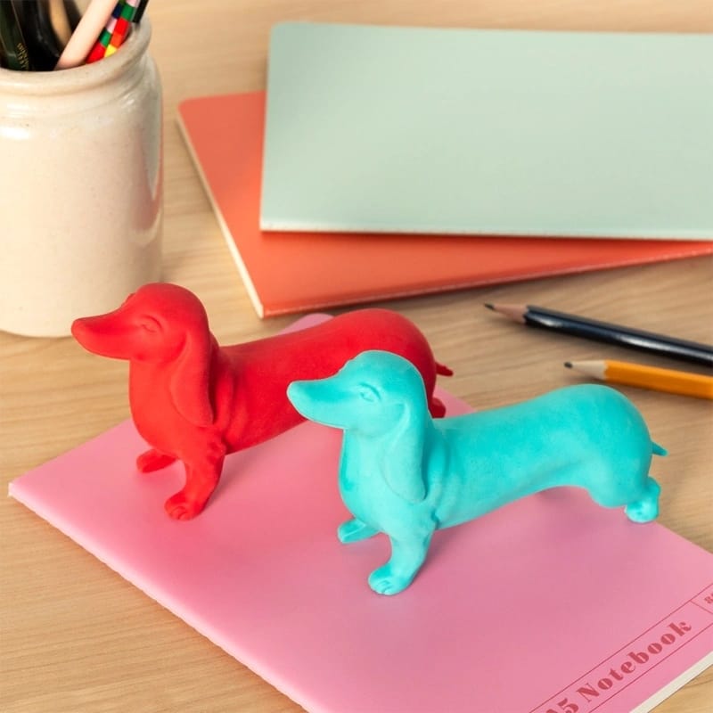 Apontadores de lápis em forma de cão dachshund vermelho e azul sobre um caderno rosa numa mesa de madeira