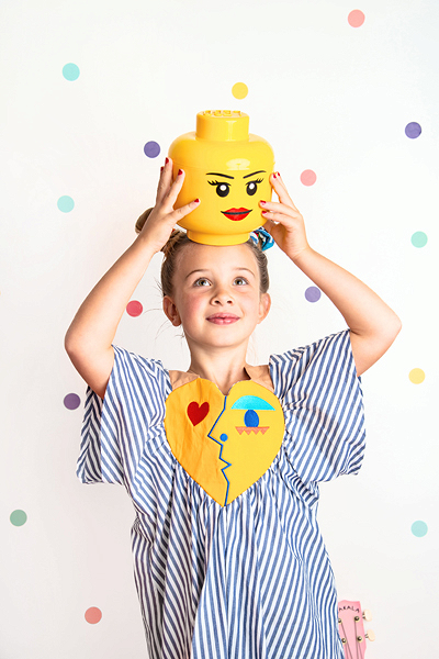 Menina com vestido riscado azul e branco a segurar na cabeça balde amarelo em forma de cabeça com rosto de boneca, fundo branco com pontos coloridos.