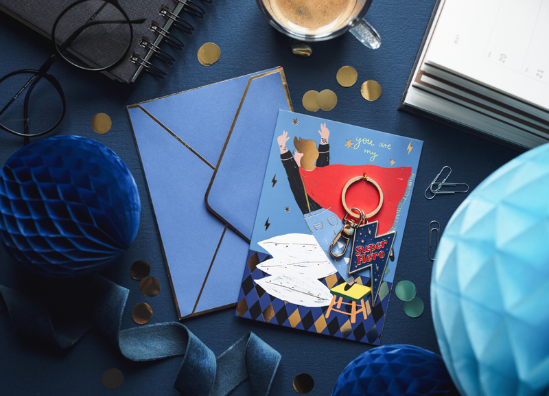 Cartão com chaveiro, envelope azul, óculos, caderno, chávena de café sobre fundo azul