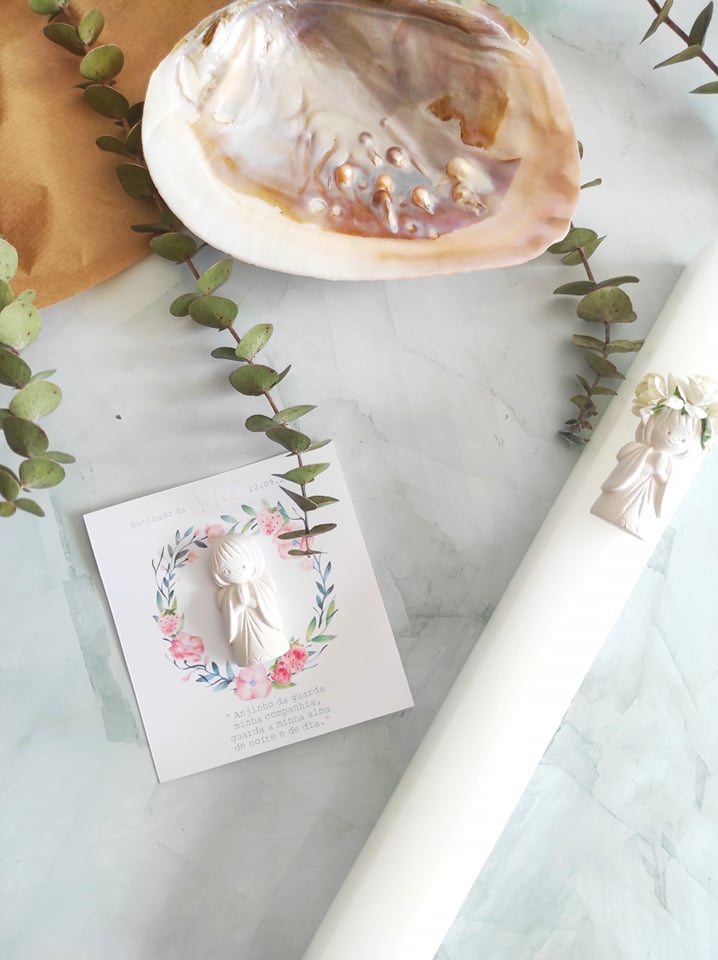 Vela decorativa branca com figura angelical, cartão florido, concha e ramos verdes