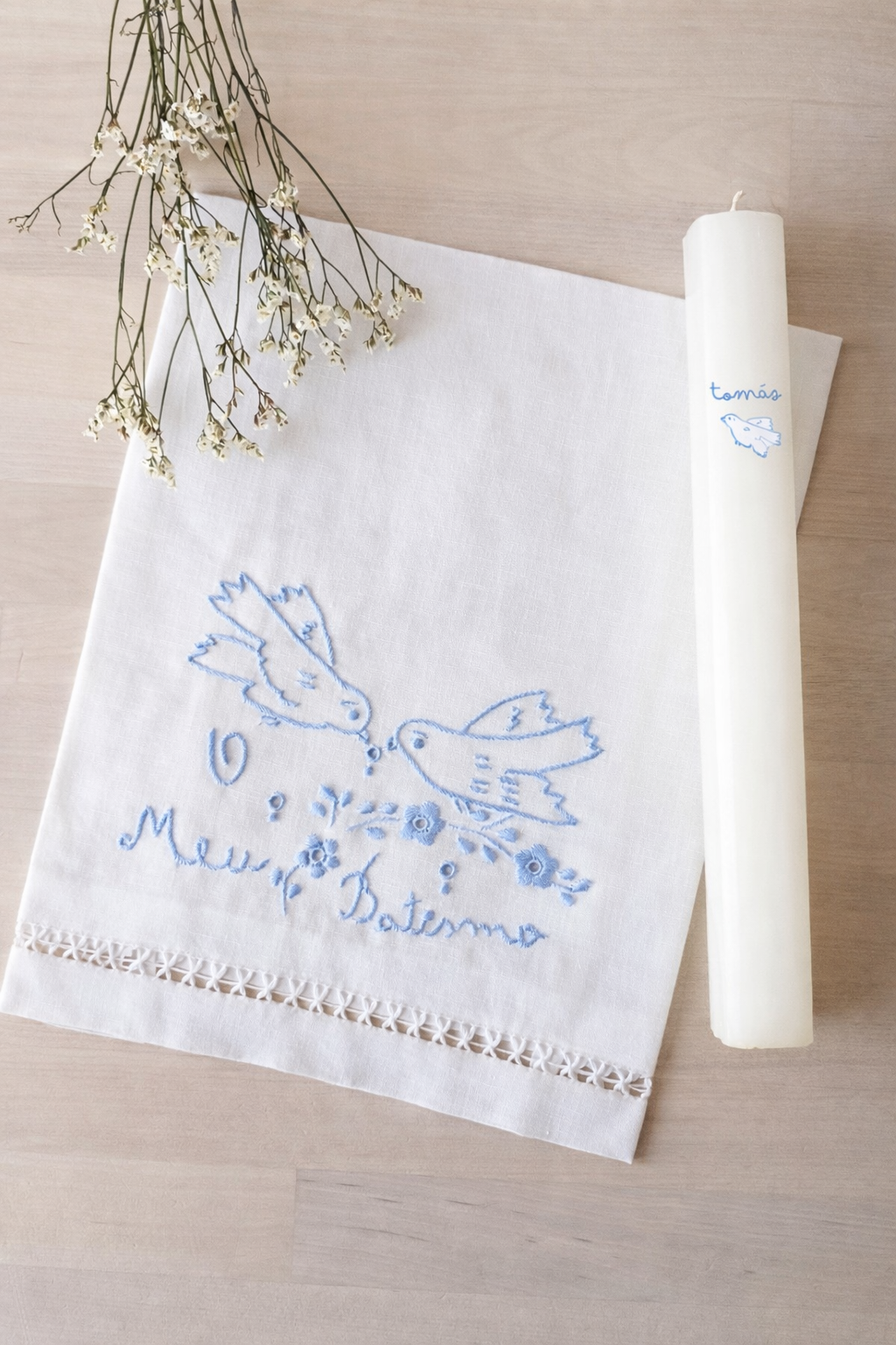 Toalha bordada branca com padrão de pássaros e vela branca decorada, em madeira clara.