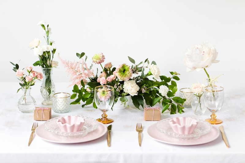 Mesa posta elegante com pratos rosa, copos de pé âmbar, talheres dourados e arranjos florais delicados em tons rosa e branco