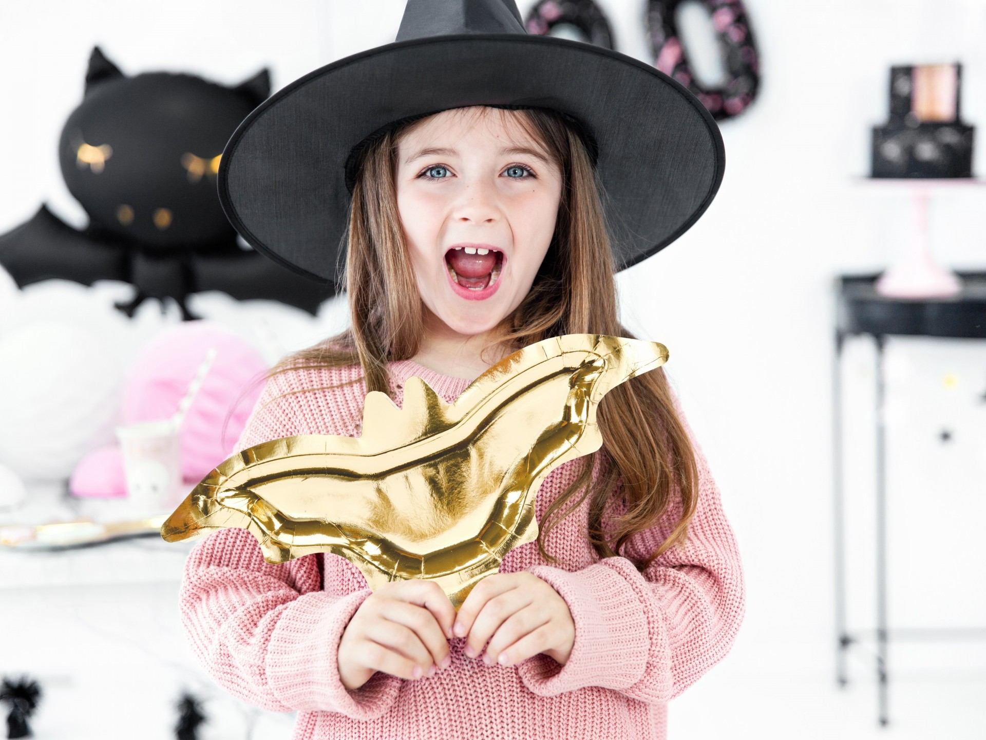 Menina segurando balão dourado em forma de morcego com decoração de Halloween ao fundo