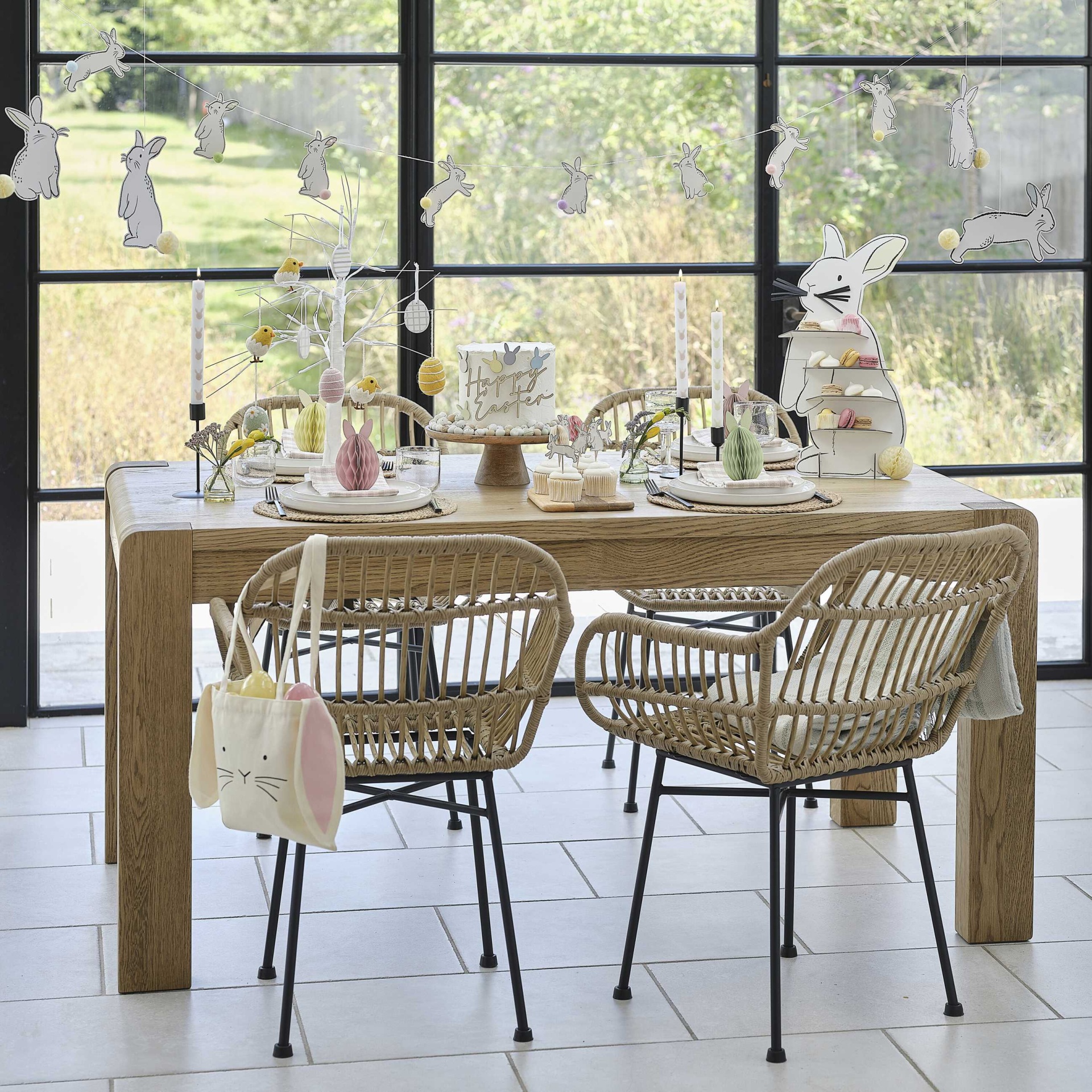 Mesa decorada para Páscoa com bolo, cadeiras de palha sintética e decoração de coelhos.