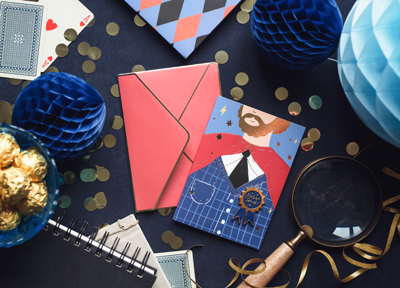 Itens de decoração e baralho em superfície escura com bolas de papel azul, envelopes vermelhos e bombons dourados