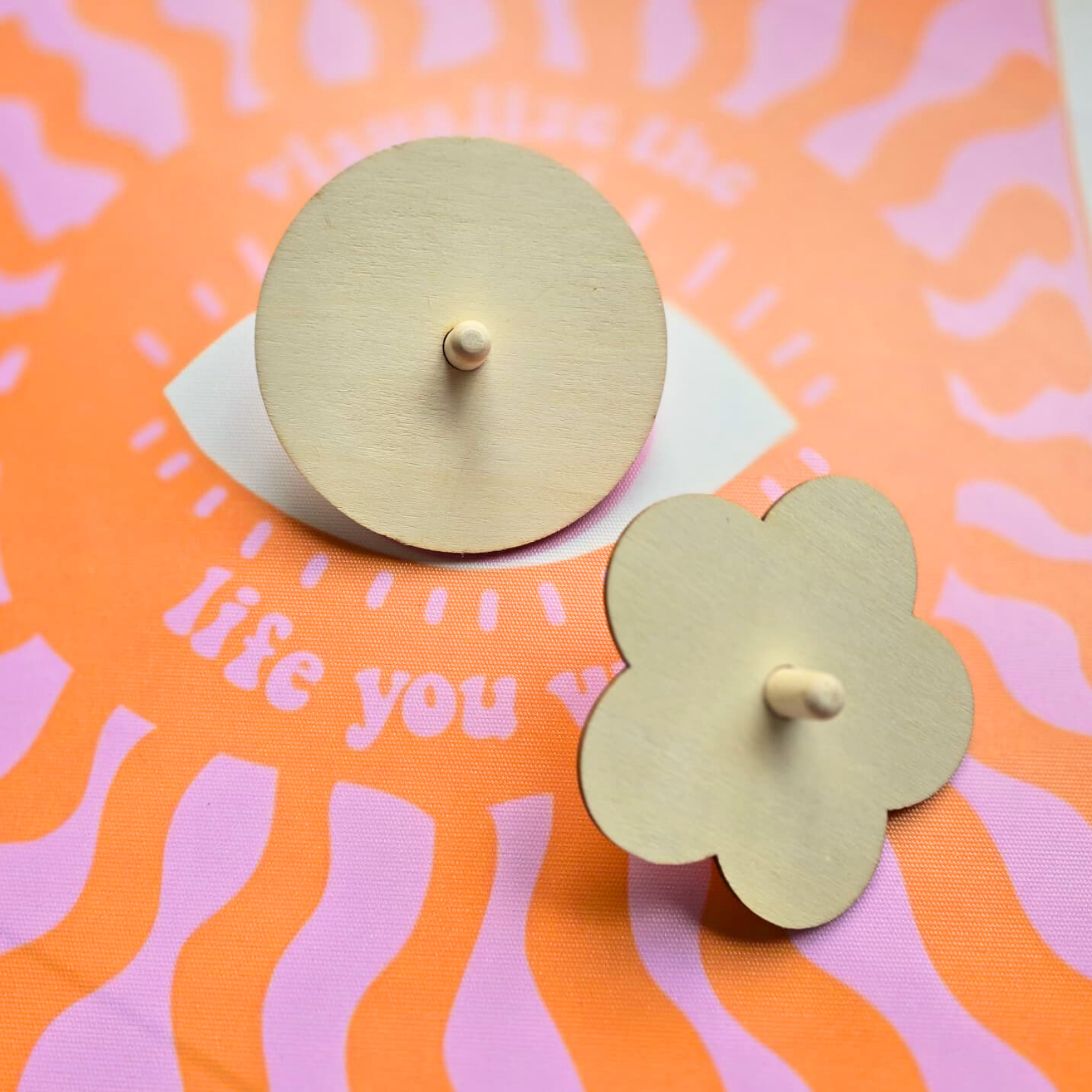 Dois suportes de vela em madeira clara, redondo e flor, sobre tecido laranja com padrão rosa e texto parcial.