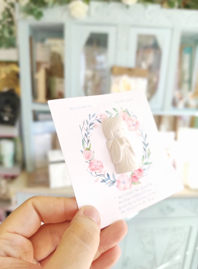 Cartão decorativo com anjo em relevo e ornamento floral nas mãos de uma pessoa, interior desfocado ao fundo