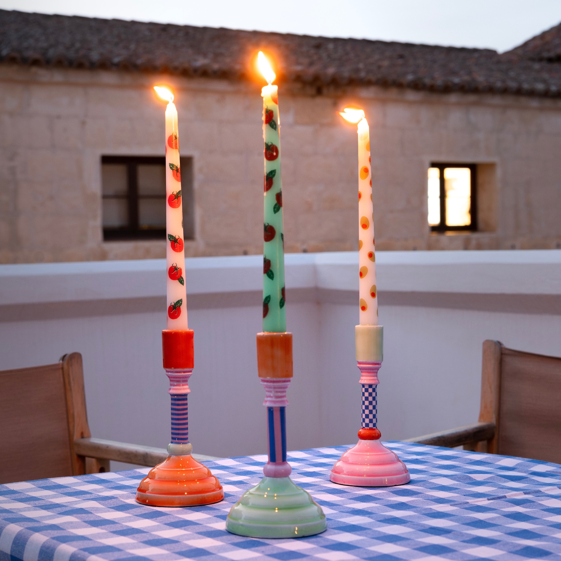 Três velas coloridas em suportes de cerâmica numa mesa com toalha às quadrículas ao ar livre