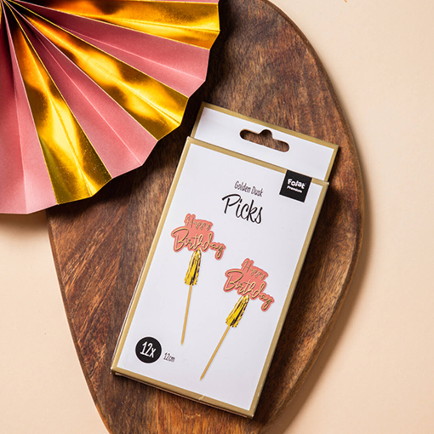 Toppers de bolo 'Happy Birthday' rosa e dourado em embalagem branca sobre tábua de madeira escura com decoração rosa e dourada ao lado.
