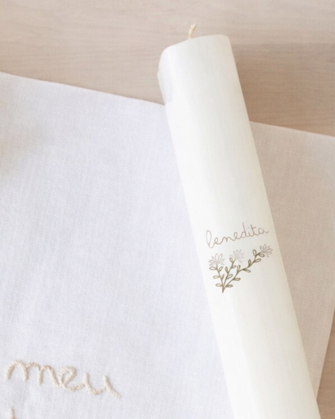 Vela branca cilíndrica com ilustração floral e texto benedita ao lado de pano branco com bordado