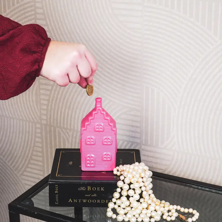 Mealheiro rosa em forma de casa com moeda, sobre livros e colar de pérolas, fundo cinza texturado.