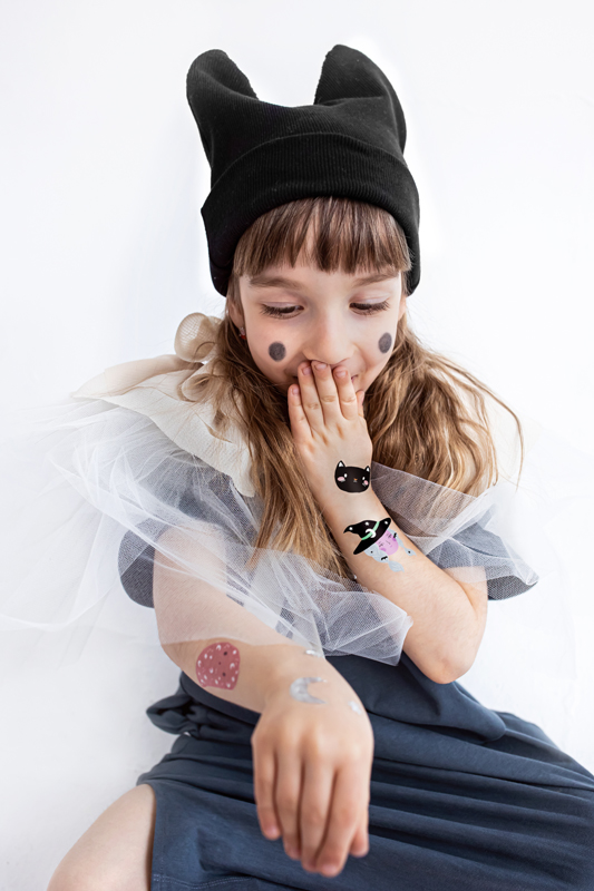 Criança a usar gorro preto com orelhas, vestido cinza com tule branco, com tatuagens temporárias e pintas pretas nas bochechas.