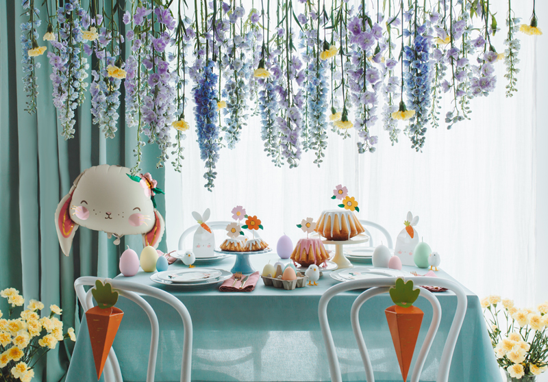 Mesa decorada com bolos, ovos e coelhos para ocasião especial com cadeiras decoradas e flores penduradas