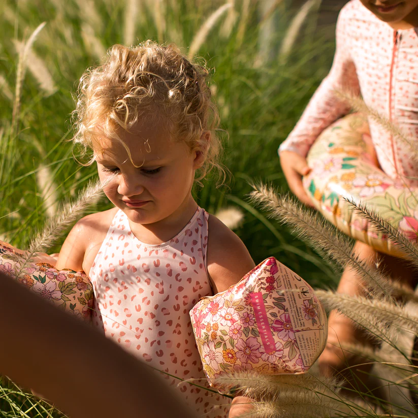 Criança loira com boias de braço florais e fato de banho cor-de-rosa num campo de relva alta