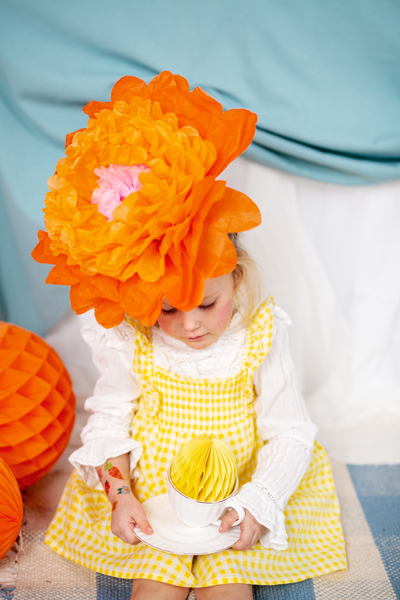 Criança com chapéu de flor laranja segurando chávena branca com objeto de papel amarelo sentada em tapete, fundo azul e branco