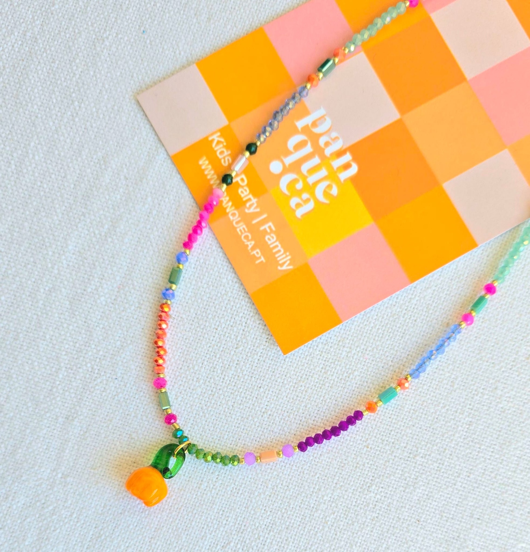 Colar colorido de contas com pendente de flor laranja e conta verde sobre cartão quadriculado com texto.