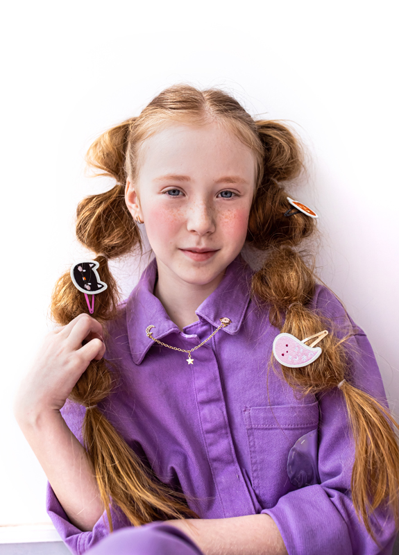 Menina com cabelo ruivo e presilhas coloridas, veste casaco roxo e colar com estrela