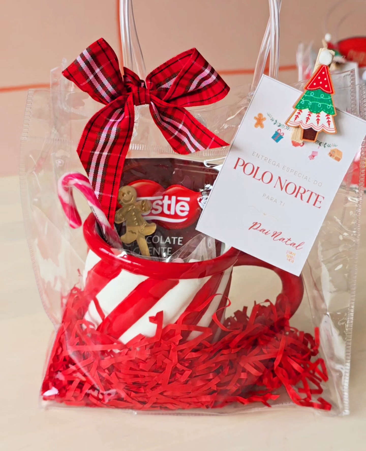 Saco transparente com caneca listrada vermelha e branca, chocolate Nestlé e cartão de Natal com laço vermelho.