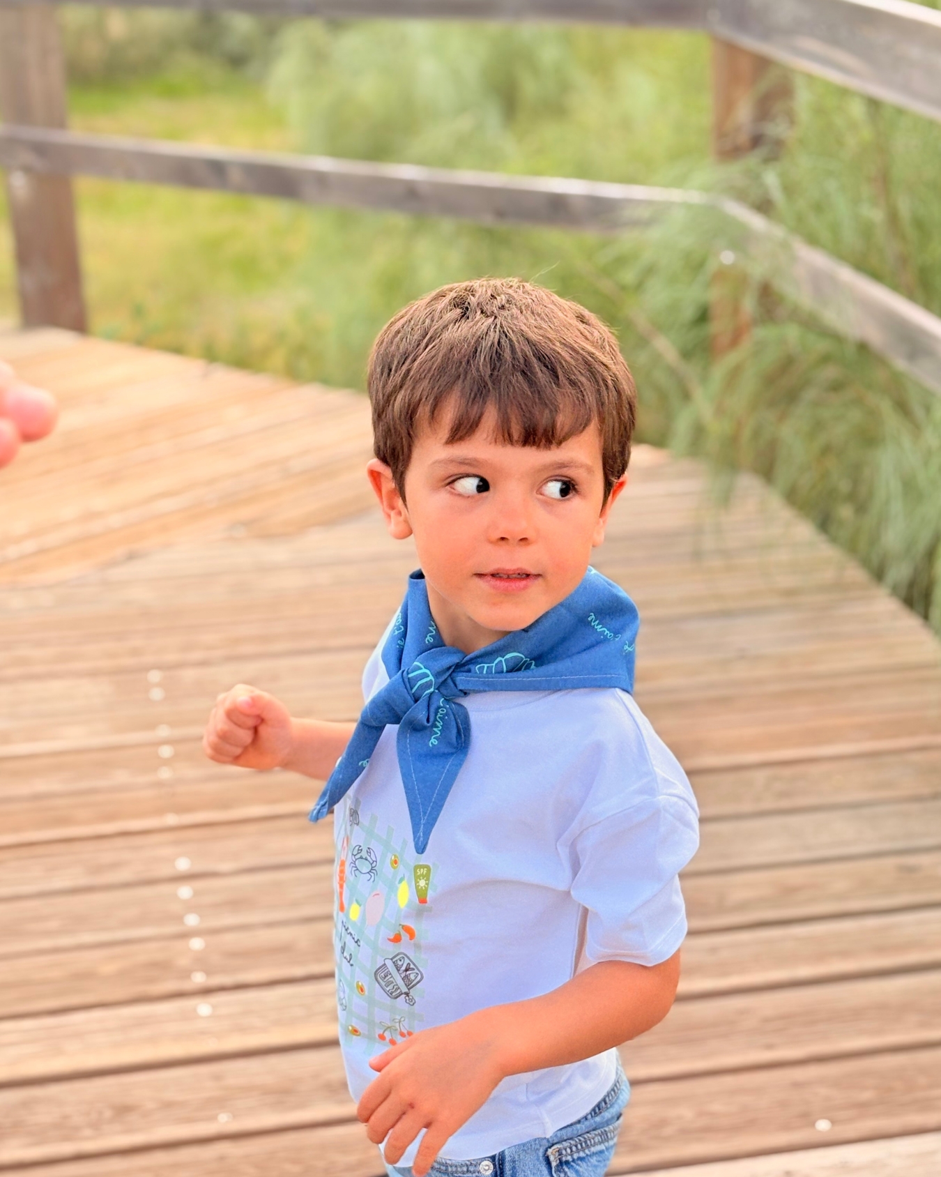 Camisola branca infantil com estampado colorido e cachecol azul numa criança em passadiço de madeira