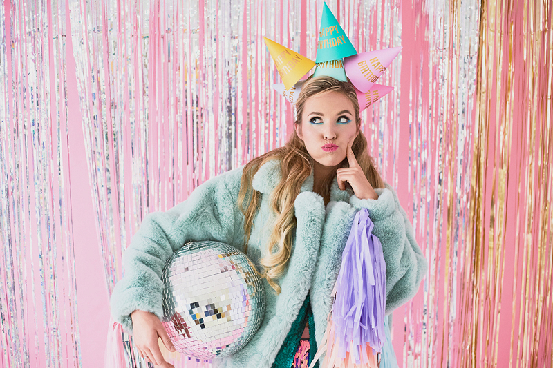 Mulher com casaco verde e chapéus de festa coloridos segurando disco espelhado e pompom lilás em fundo rosa metálico