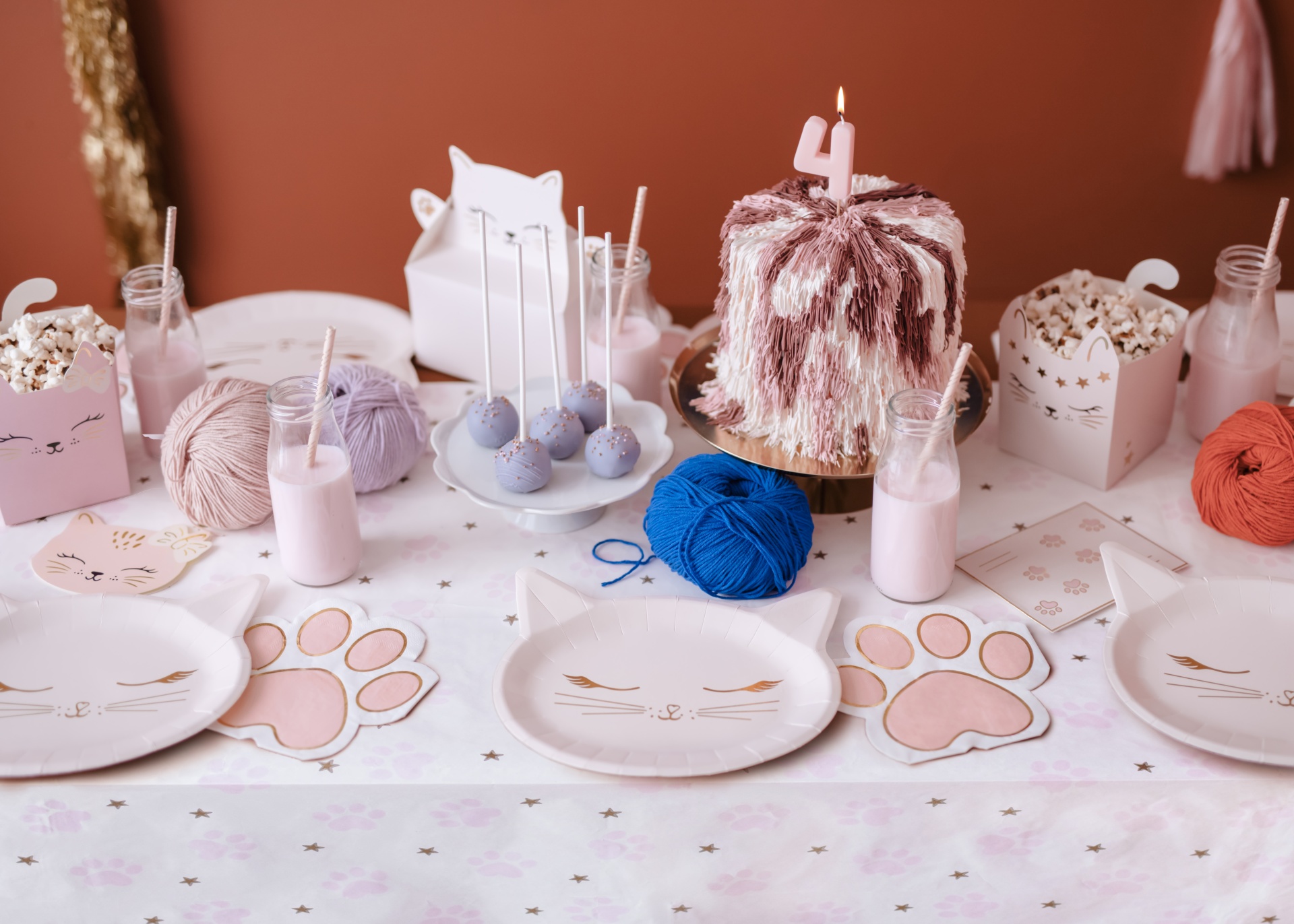 Mesa decorada para festa com tema de gatos, bolo com vela 4, pratos e guardanapos em forma de gato e pata, e bolas de lã coloridas