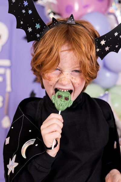 Criança ruiva com fantasia preta e máscara de morcego segura pirulito verde de monstro