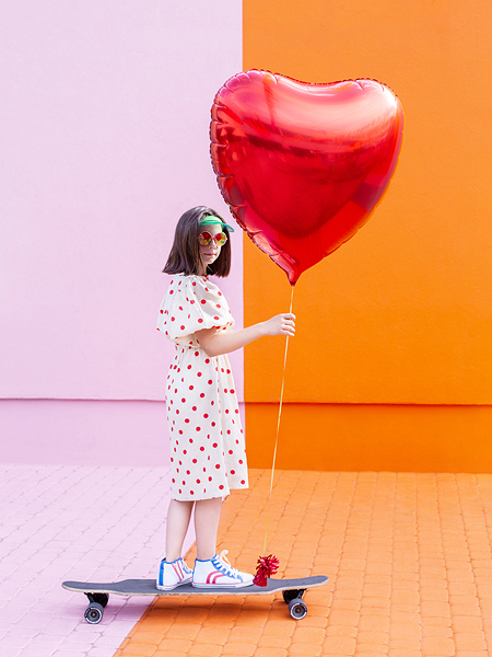 Menina com vestido branco com bolinhas vermelhas segurando balão vermelho em forma de coração em skate preto sobre fundo rosa e laranja