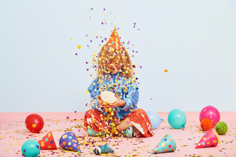 Criança com chapéu de festa rodeada por confetes e balões no chão