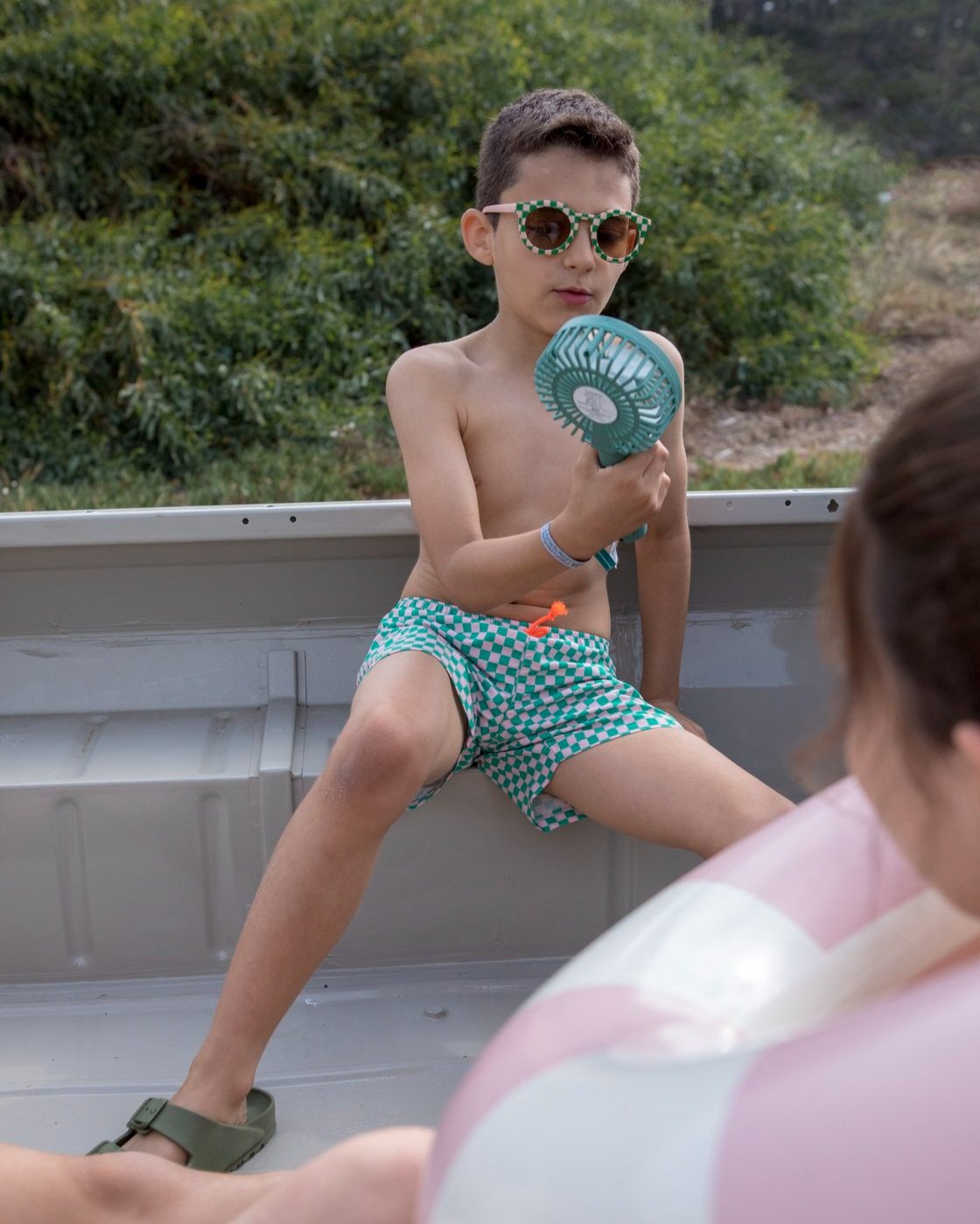 Criança de óculos de sol e calções xadrez verdes sentada numa embarcação segurando ventilador portátil verde
