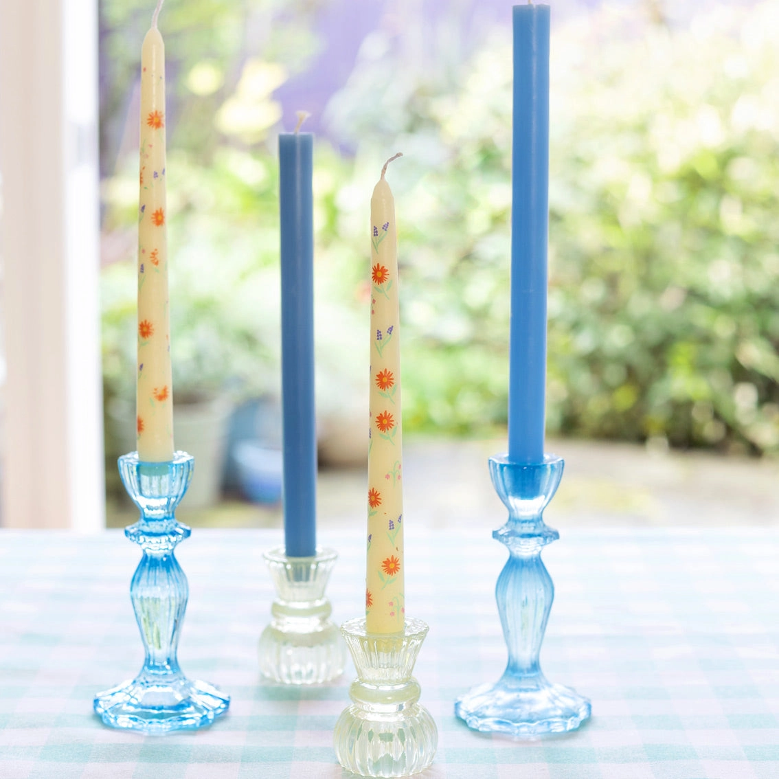 Velas decorativas em castiçais de vidro sobre mesa xadrez azul e branco com fundo de jardim desfocado