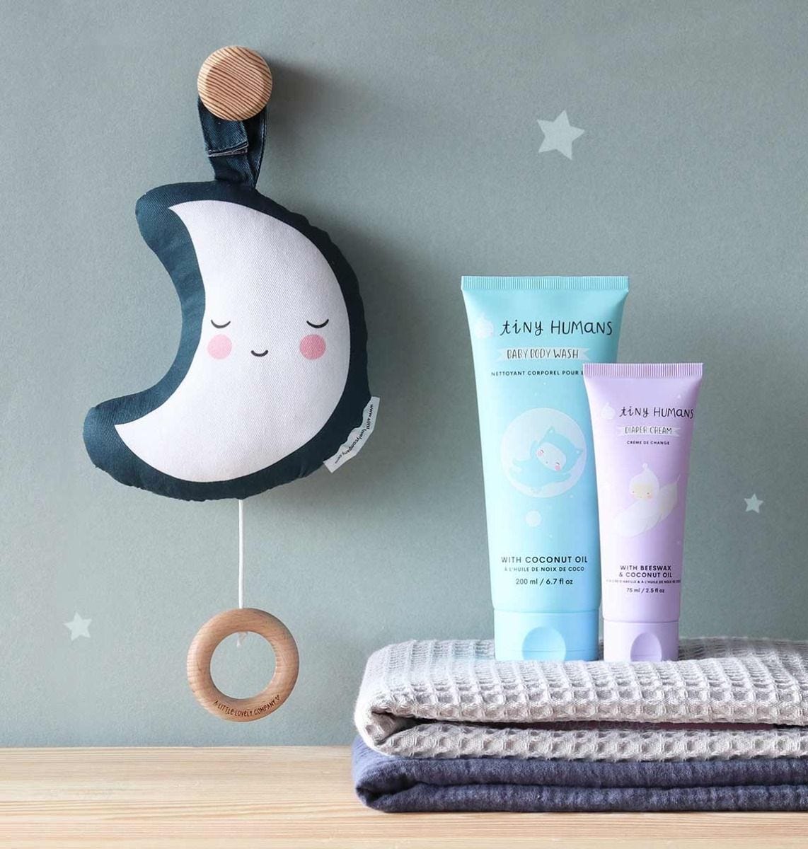 Móbile de lua em tecido azul e branco, cremes para pele em tubos azul e roxo, e toalhas cinza e bege sobre madeira