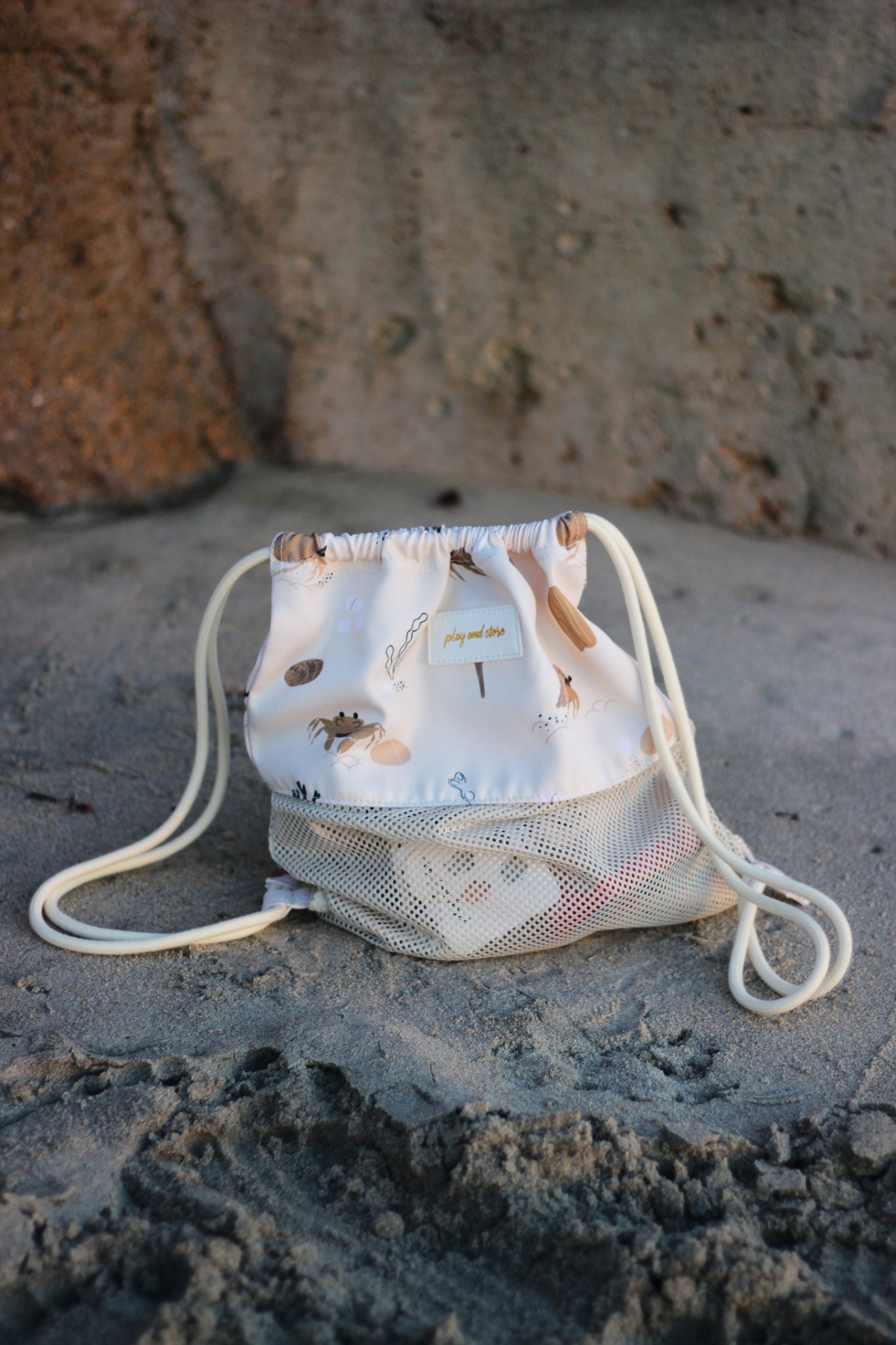 Mochila pequena com padrão floral castanho e bolsos em rede transparente numa superfície arenosa