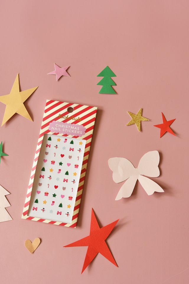Pacote de autocolantes para unhas de Natal numa superfície cor de rosa com recortes de papel colorido ao redor.