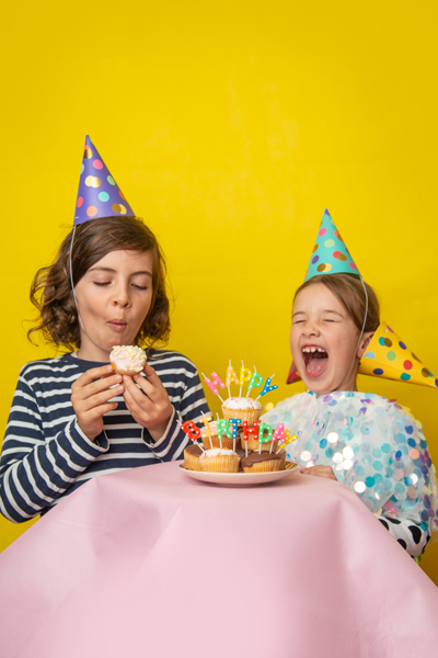 Crianças com chapéus de festa e cupcakes com velas coloridas numa mesa com toalha rosa