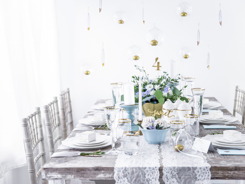 Mesa de jantar decorada com louça branca, arranjos florais azul e branco, toalha de renda e elementos em vidro suspensos