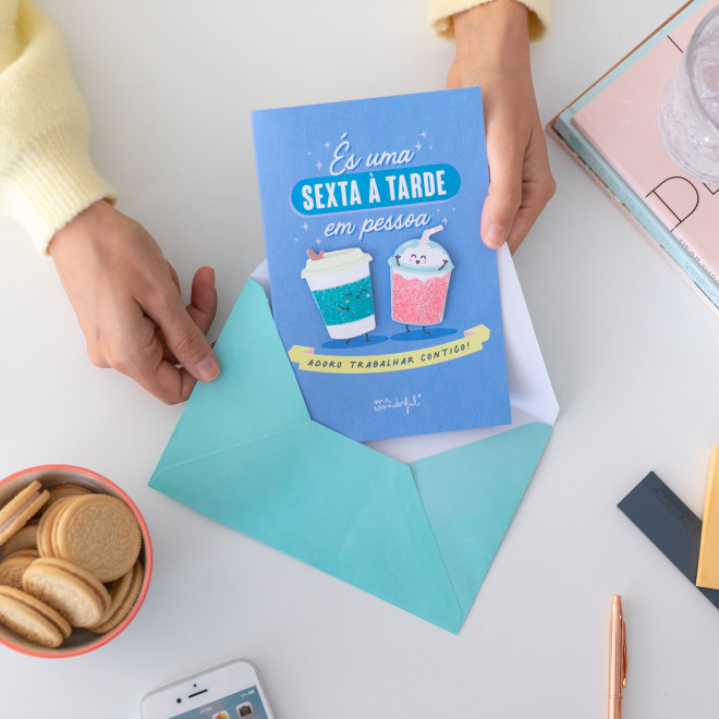 Cartão azul com texto e ilustrações de bebidas feliz sobre envelope azul claro e mesa branca com mãos, bolachas e outros objetos